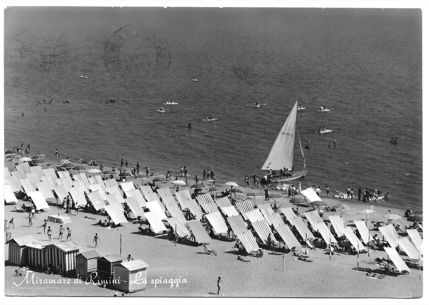 Zwart-wit ansichtkaart van het strand van Rimini rond 1960, met rijen parasols en een zeilboot.