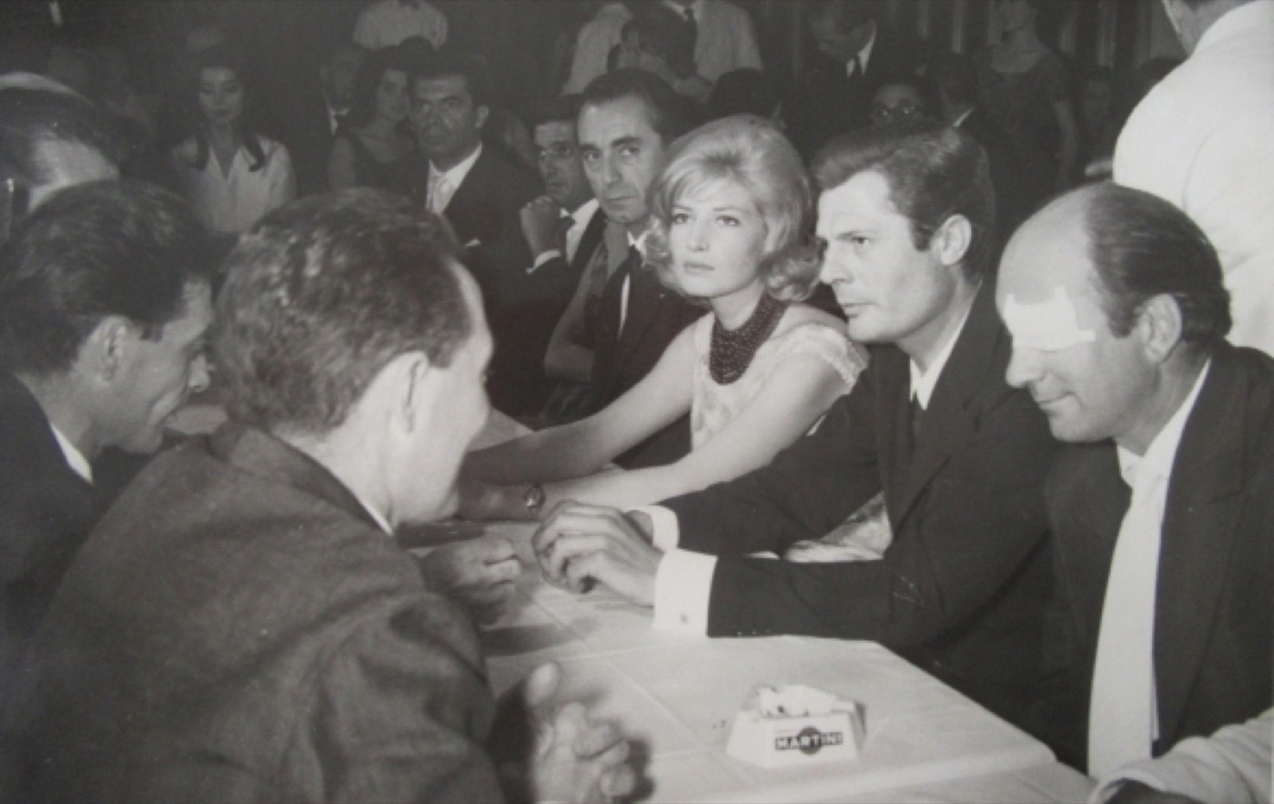 Zwart-witfoto uit 1960: gasten aan een gedekte tafel, in gesprek.