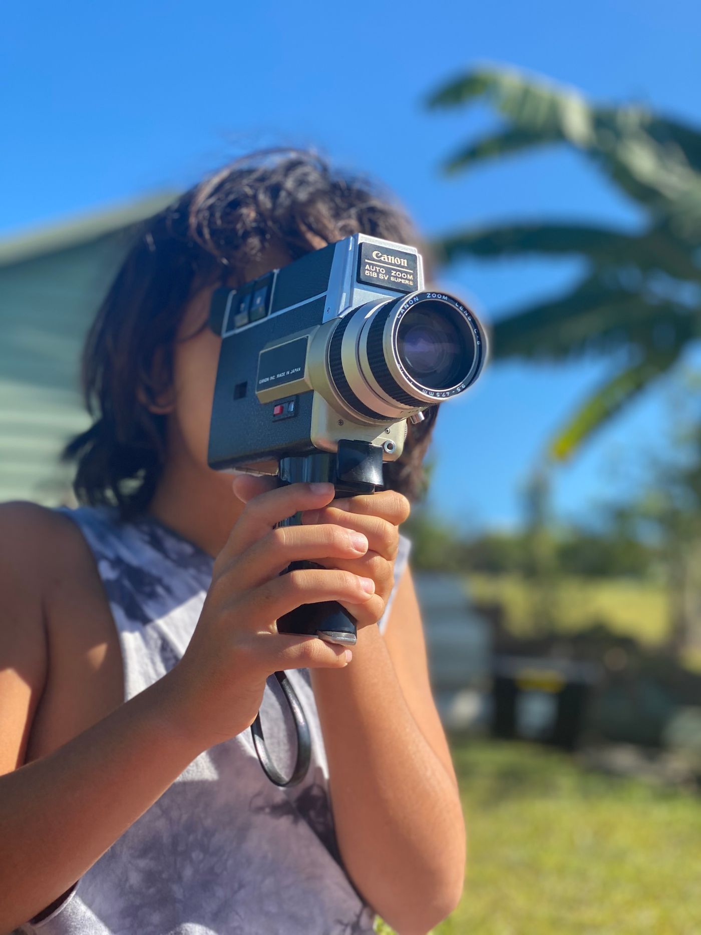 Een vrouw filmt met een oude filmcamera, met palmbomen en blauwe lucht achter haar.